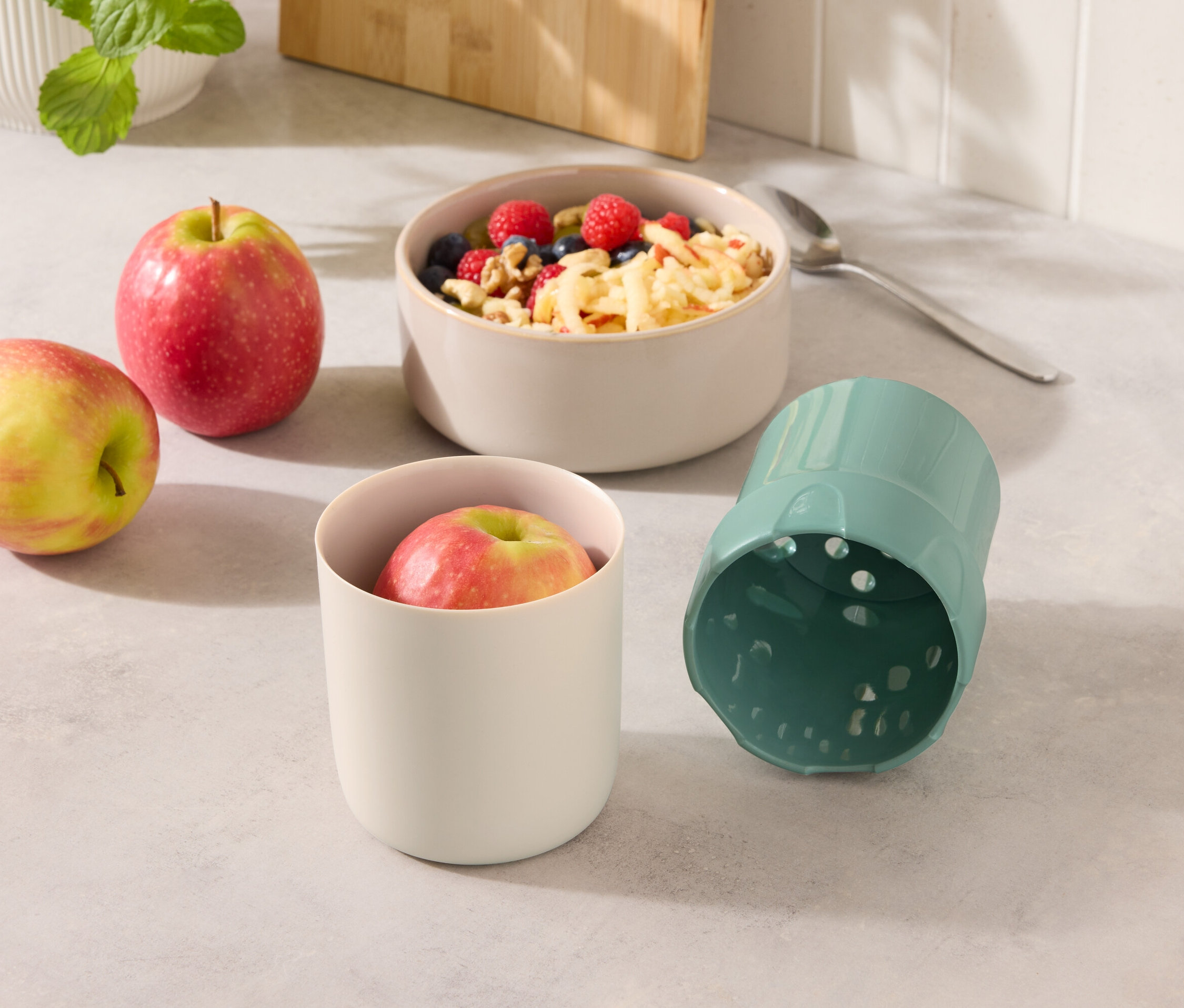 Zwei Äpfel neben einer Müslischale und einem Becher mit Apfel und einem blauen Becher.