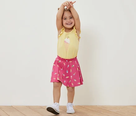 Lächelndes Mädchen steht mit erhobenen Händen und trägt ein gelbes T-Shirt und einen Kinder-Jerseyrock mit Hose, Eis.