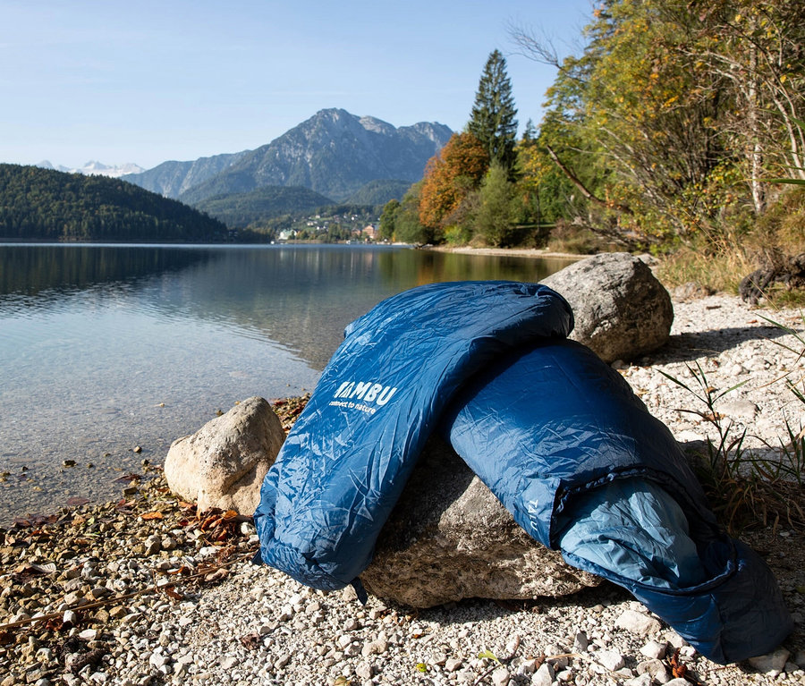 Blauer Tambu Mumienschlafsack »TALU 1500« liegt auf einem Stein an einem Kiesstrand am See.