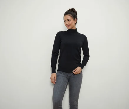 Frau mit braunen Haaren zum Dutt hochgesteckt posiert in schwarzem Rollkragenpullover und grauen Jeans.