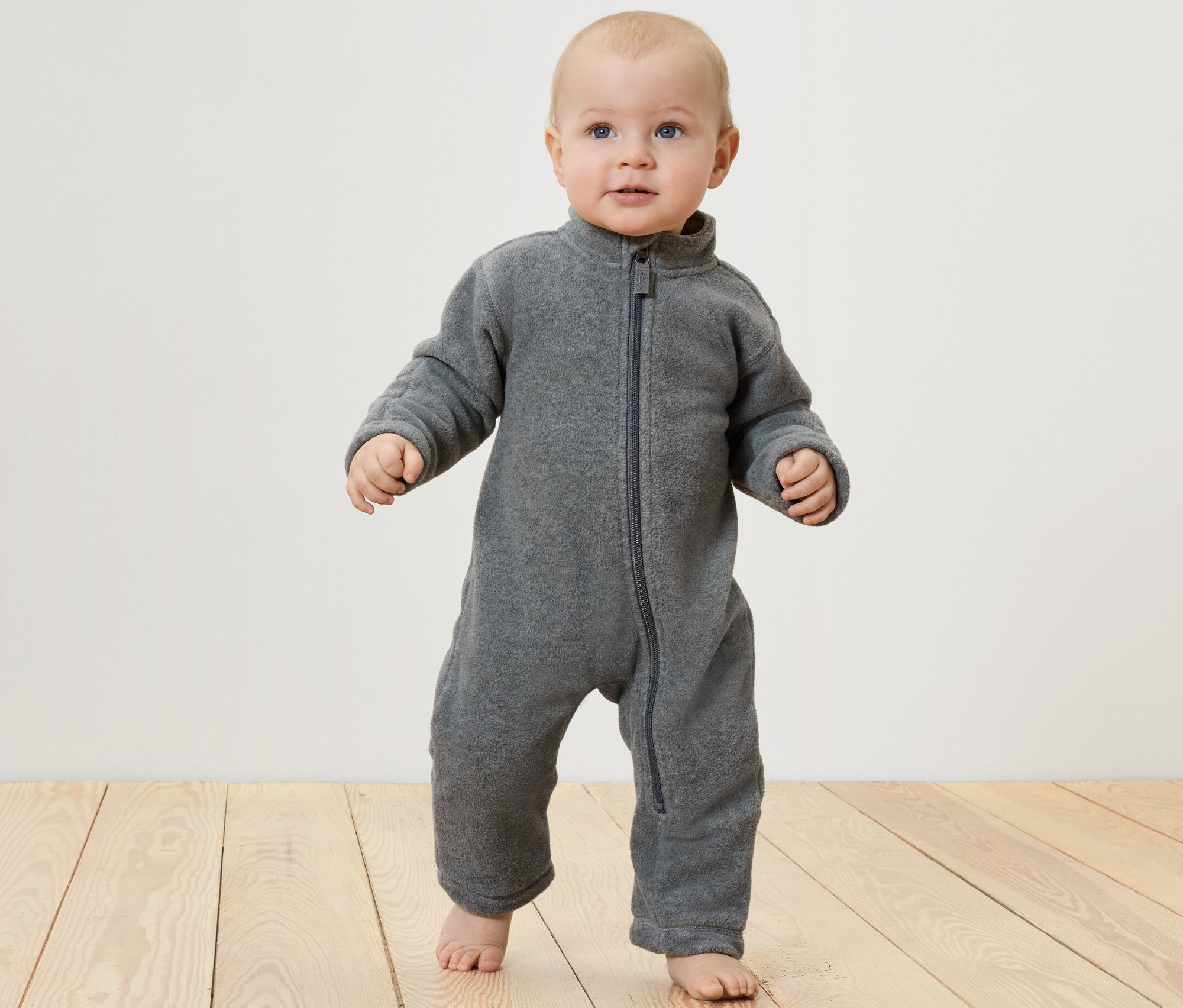 Baby steht auf einem Holzboden und trägt einen Baby-Fleece-Einteiler.