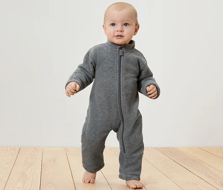 Baby steht auf einem Holzboden und trägt einen Baby-Fleece-Einteiler.