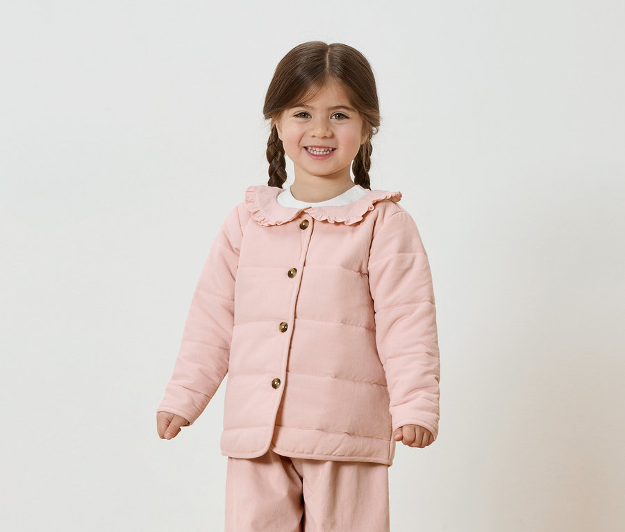 Mädchen mit Zöpfen trägt eine rosa Jacke und Hose vor einem weißen Hintergrund.