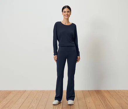 Frau mit dunklem Haar, die ein marineblaues, langärmeliges Oberteil und eine passende Hose vor einer weißen Wand trägt.