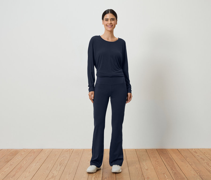 Frau mit dunklem Haar, die ein marineblaues, langärmeliges Oberteil und eine passende Hose vor einer weißen Wand trägt.