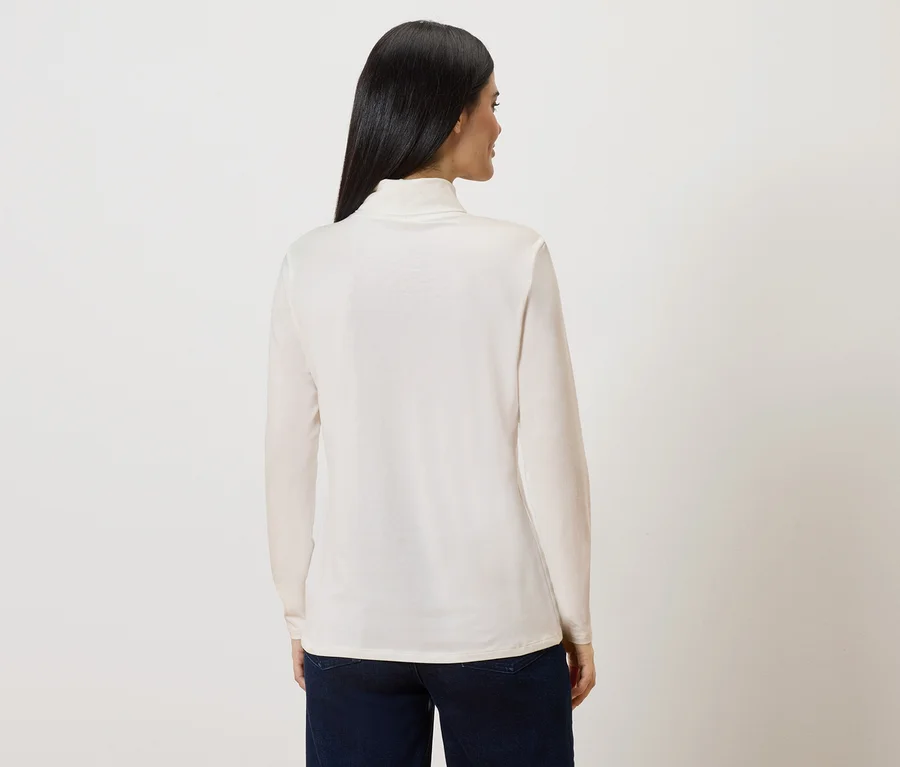 Rückansicht einer Frau mit langen, dunklen Haaren, die ein cremefarbenes Rollkragenshirt und dunkle Jeans trägt.