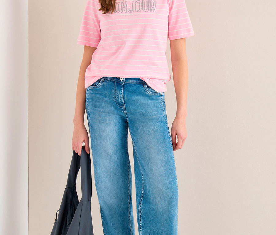 Lächelnde Frau trägt ein pink gestreiftes CECIL Shirt mit „BONJOUR“-Aufschrift, blaue weite Jeans und silberne Sneaker, eine dunkelgraue Umhängetasche haltend.