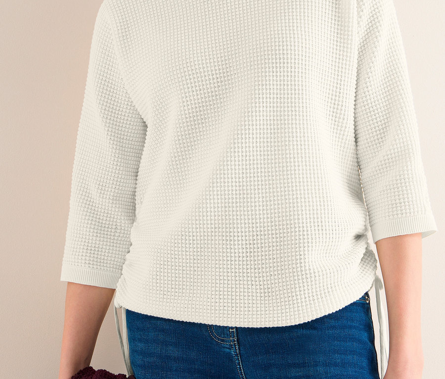 Frau trägt einen cremefarbenen CECIL Feinstrickpullover mit 3/4-Ärmeln und seitlichem Kordelzug, blaue Jeans, ein bordeauxfarbenes Kleidungsstück haltend.