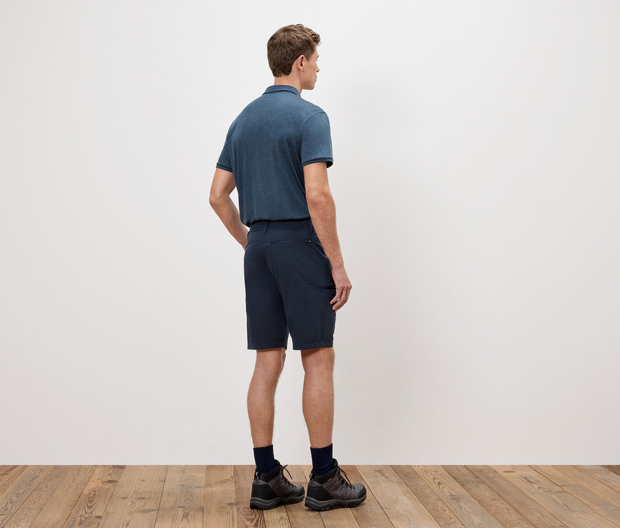 Ein Mann steht mit dem Rücken zur Kamera, trägt ein blaues Poloshirt, Shorts und Wanderschuhe vor einer weißen Wand.