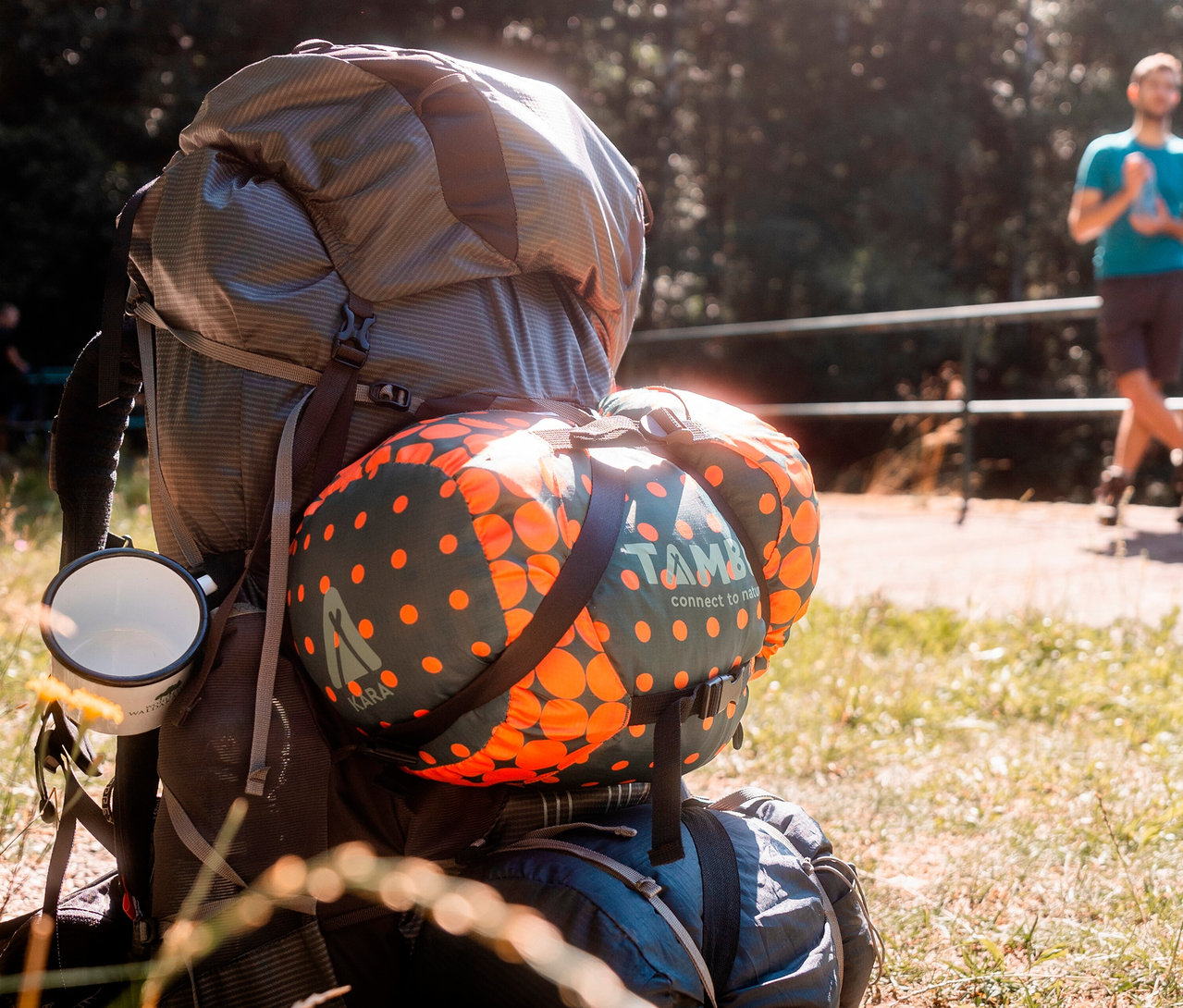 Rucksack mit befestigtem, orange gepunktetem Tambu »KARA 1.600« Mumienschlafsack. Im Hintergrund geht eine Person auf einem Weg.
