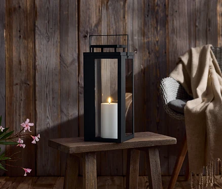 Eine schwarze Laterne mit einer weißen Kerze steht auf einem kleinen Holztisch. Im Hintergrund befindet sich eine Holzwand und ein Stuhl mit einer Decke.