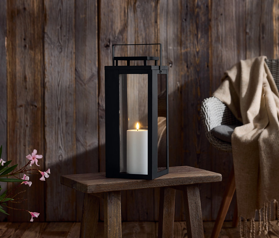 Eine schwarze Laterne mit einer weißen Kerze steht auf einem kleinen Holztisch. Im Hintergrund befindet sich eine Holzwand und ein Stuhl mit einer Decke.
