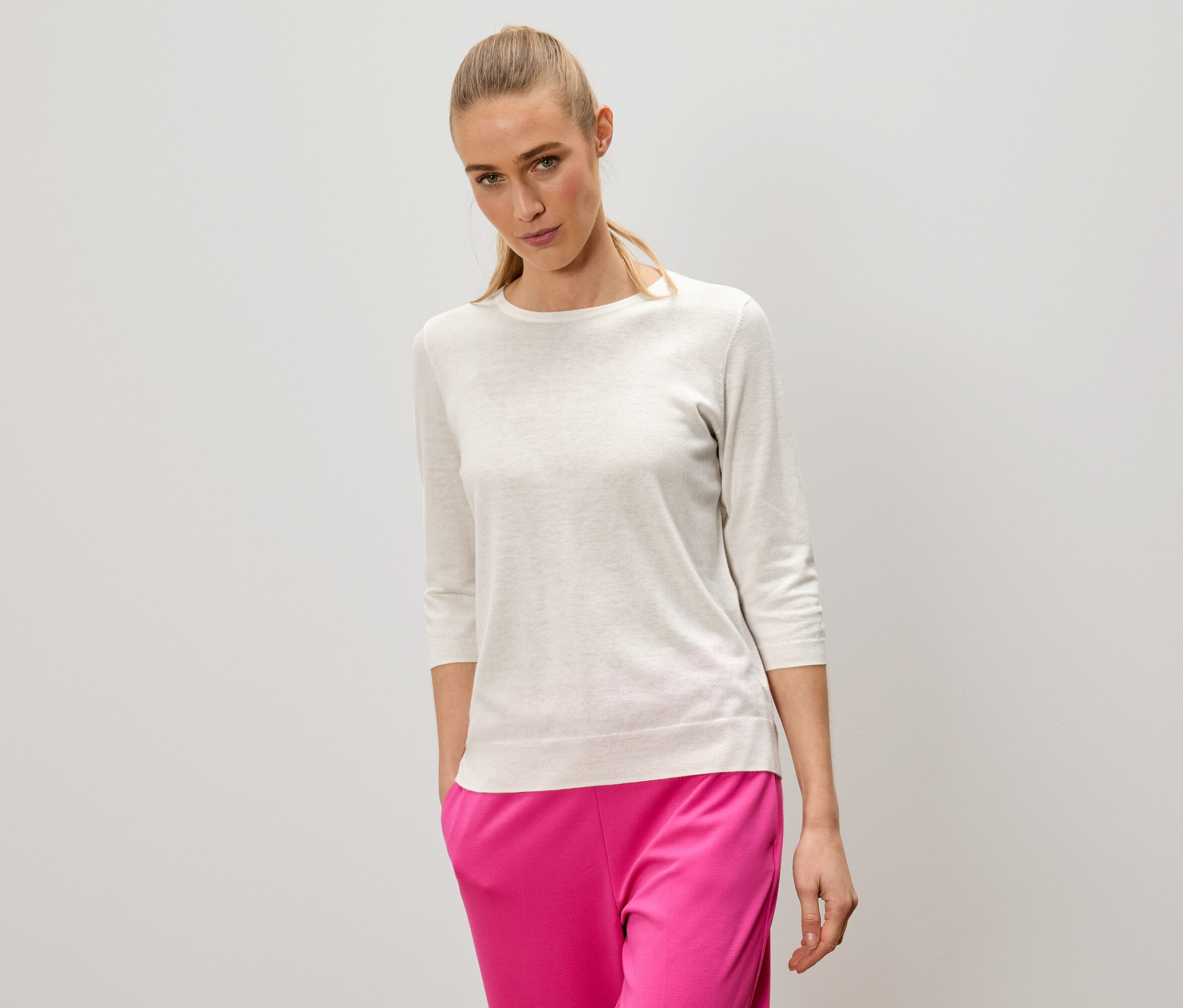 Frau mit blonden Haaren trägt einen hellen Pullover und pinke Hosen.