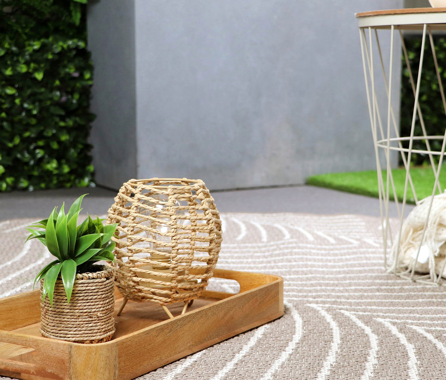 Dekorative Tischszene mit einer Pflanze in einem geflochtenen Topf und einer Lampe auf einem Holztablett, im Hintergrund ein Beistelltisch.