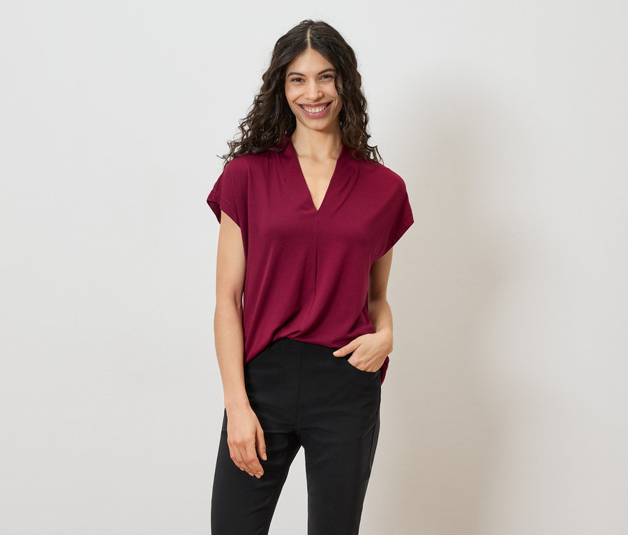Frau mit lockigem, dunklem Haar trägt ein weinrotes T-Shirt und schwarze Hosen vor einem weißen Hintergrund.