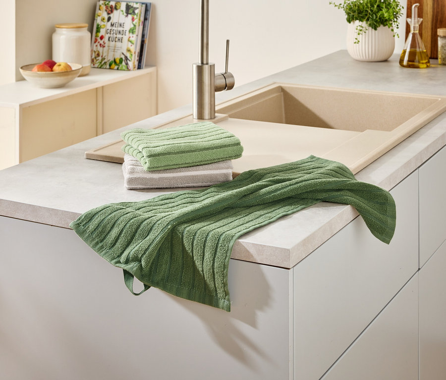 Auf einer Küchentheke liegen ein Stapel und ein einzelnes grünes Geschirrtuch neben einer Spüle und einem Wasserhahn.