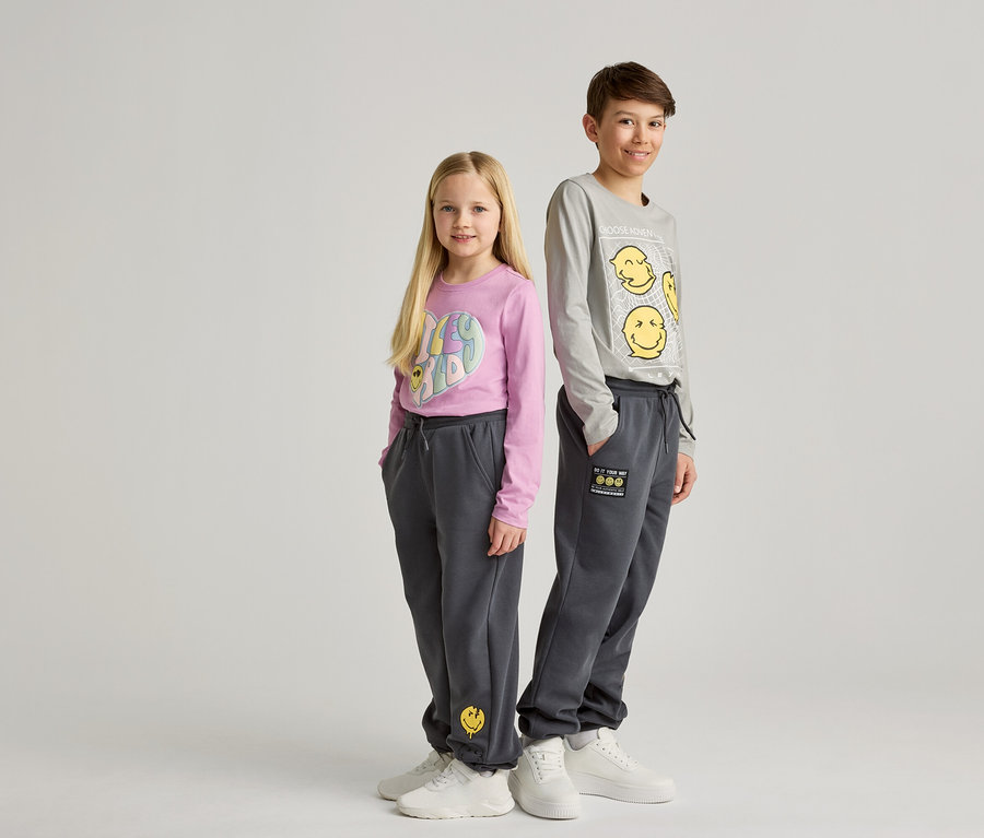 Ein Mädchen mit einem rosa Hemd und ein Junge mit einem grauen Hemd stehen nebeneinander und tragen graue Jogginghosen und weiße Turnschuhe.