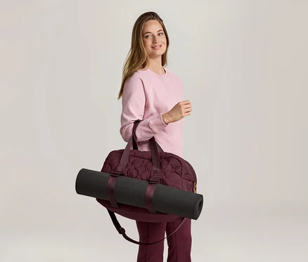 Frau mit rosa Pullover und weinroter Hose, die eine weinrote Sporttasche mit einer schwarzen Yogamatte über der Schulter trägt.