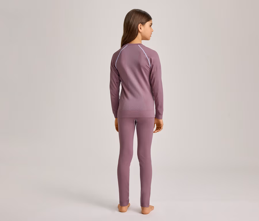 Mädchen von hinten, das ein langärmeliges, lilafarbenes Oberteil mit Muster und passende Leggings trägt. Sie steht barfuß vor einem hellen Hintergrund.