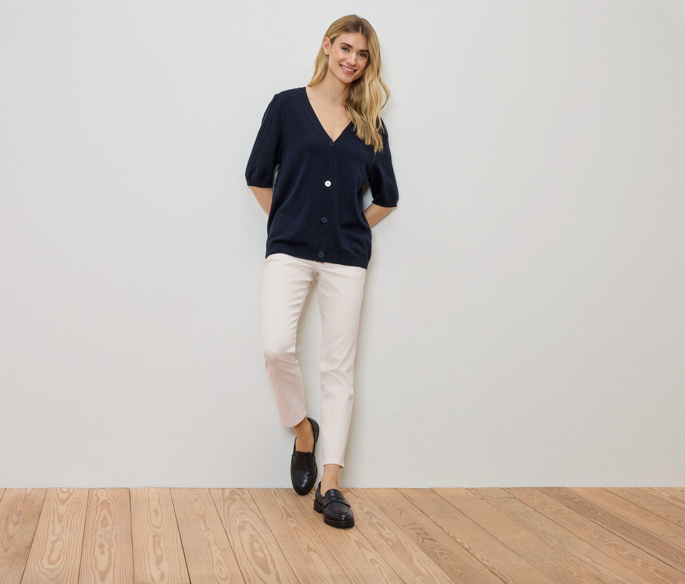 Frau steht an einer Wand, trägt eine marineblaue Strickjacke, eine helle Hose und schwarze Schuhe.