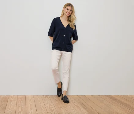 Frau steht an einer Wand, trägt eine marineblaue Strickjacke, eine helle Hose und schwarze Schuhe.