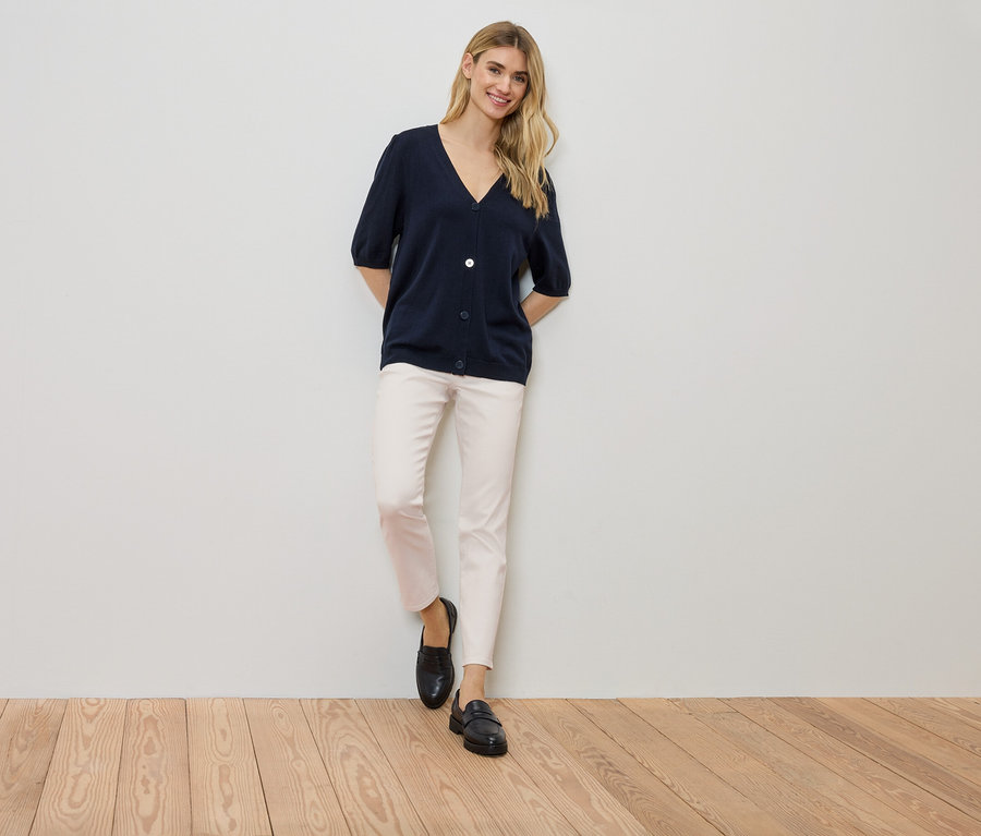 Frau steht an einer Wand, trägt eine marineblaue Strickjacke, eine helle Hose und schwarze Schuhe.