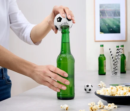 Eine Person öffnet eine grüne Bierflasche mit einem Fußball-Flaschenöffner. Auf dem Tisch befinden sich Popcorn und andere Flaschen.