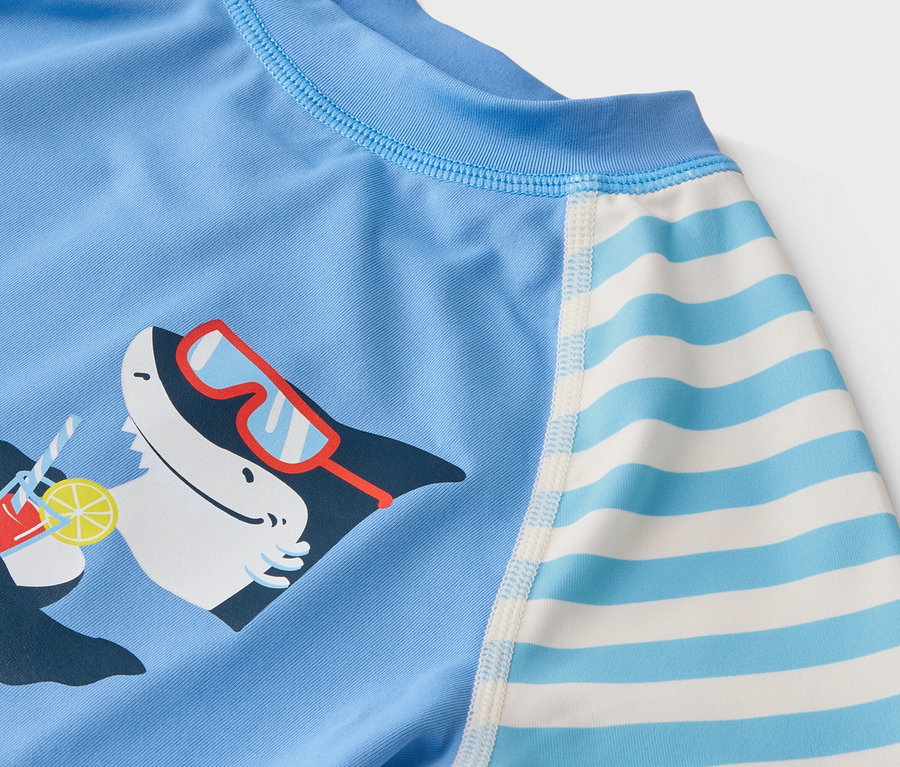 Detailaufnahme eines hellblauen Kinder-Badeoberteils mit einem Hai, der eine Sonnenbrille trägt und einen Cocktail hält, sowie gestreiften Ärmeln.