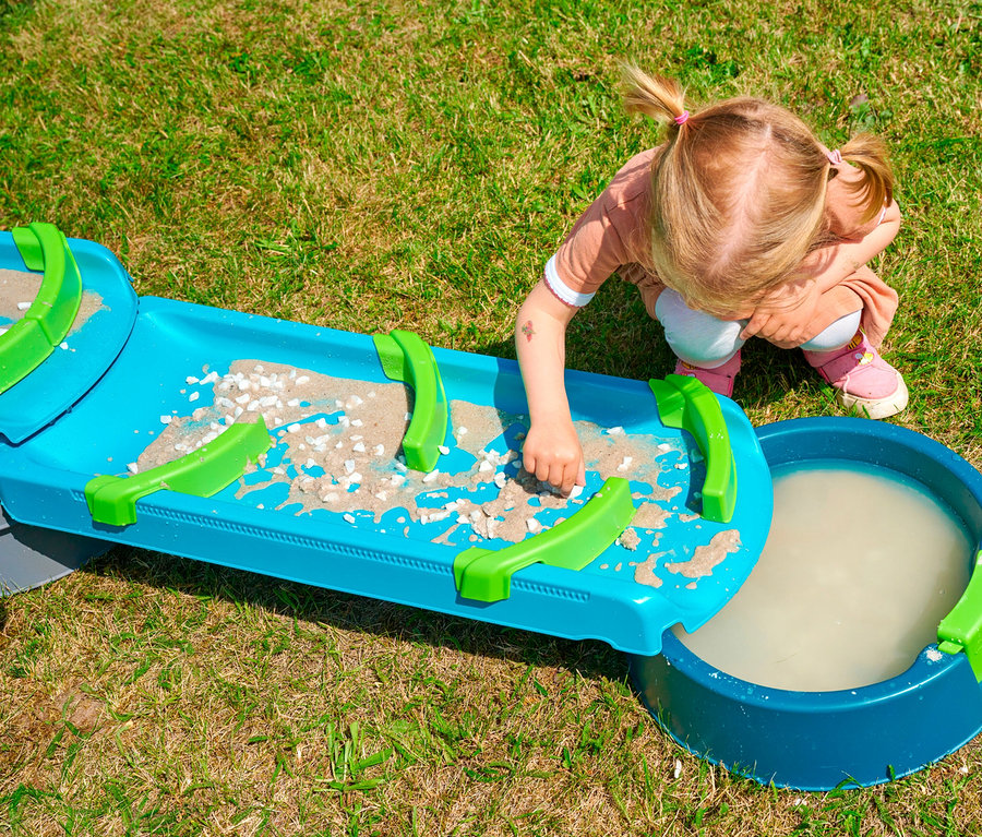 Mädchen spielt mit AquaPlay »Mudway STREAM« Matschbahn auf Rasen. Die Bahn ist blau mit grünen Elementen und Sand.