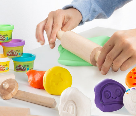 Eine Person rollt grüne Play-Doh-Knete mit einem Nudelholz aus. Mehrere Dosen Play-Doh-Knete und geformte Knete liegen auf dem Tisch.