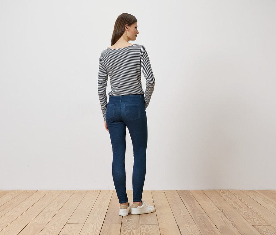 Eine Frau steht mit dem Rücken zum Betrachter, trägt Jeans, ein gestreiftes Langarmshirt und weiße Sneaker.