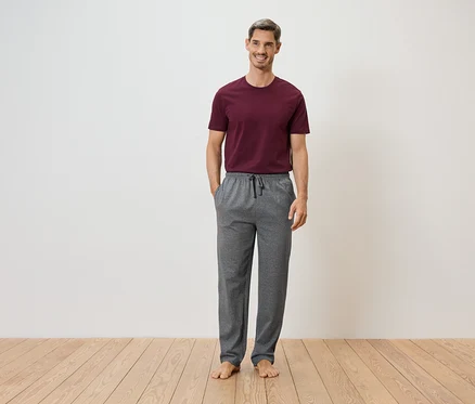 Ein Mann steht barfuß vor einer weißen Wand und einem Holzboden. Er trägt ein weinrotes T-Shirt und eine graue Jogginghose mit Kordelzug.