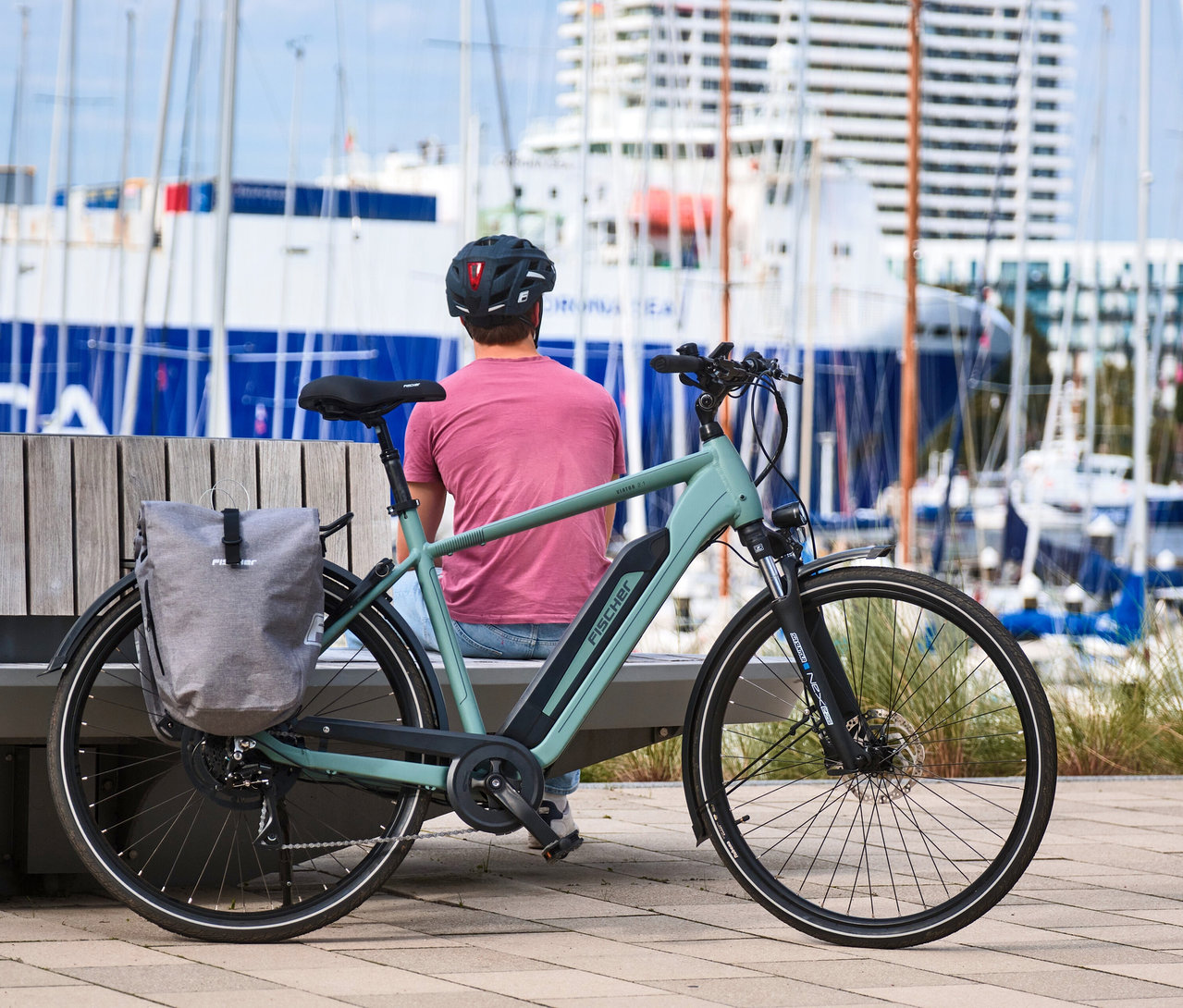 Mann sitzt auf Bank neben großem FISCHER Trekking E-Bike VIATOR 2.1 ACTIVE mit Diamant-Rahmen vor Hafen.