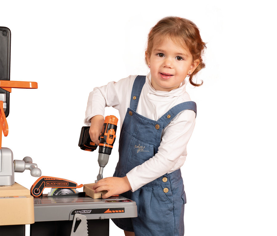 Mädchen in Jeanslatzhose bohrt mit einem Black+Decker Spielzeugbohrer in ein Stück Holz auf einer Spielzeugwerkbank.