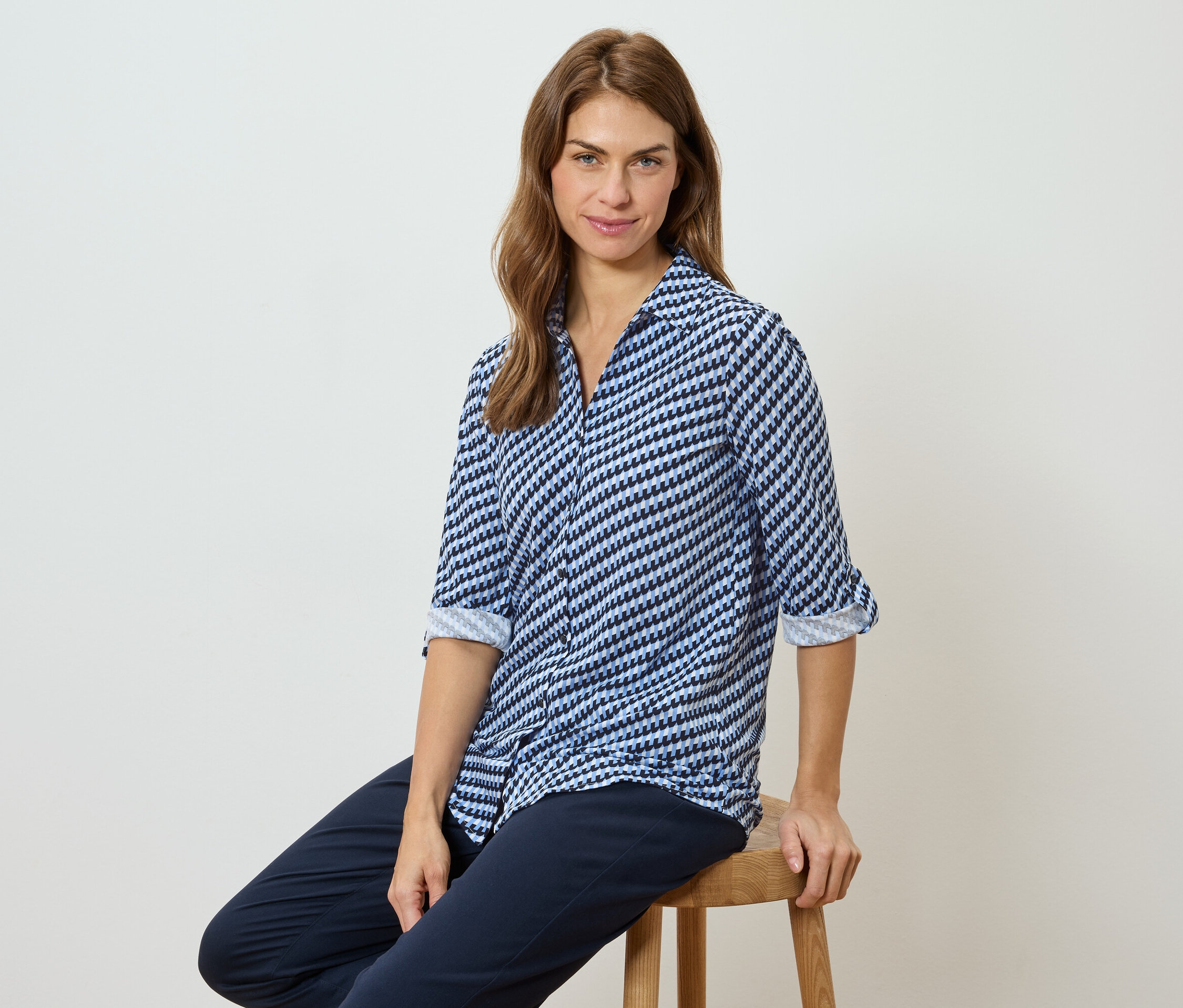 Frau sitzt auf einem Hocker und trägt eine blau-weiß gemusterte Bluse und eine dunkle Hose.