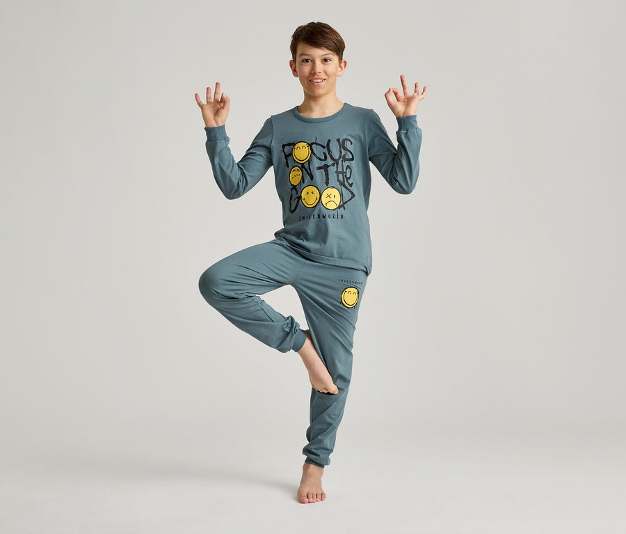 Junge in blauem Schlafanzug mit Smiley-Motiv, balanciert auf einem Bein und formt mit den Händen Mudras.