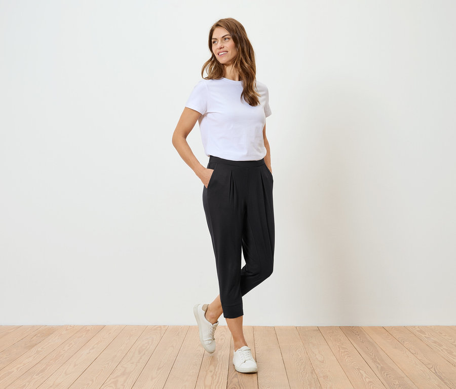Frau mit weißem T-Shirt und schwarzer Hose posiert vor einer weißen Wand.
