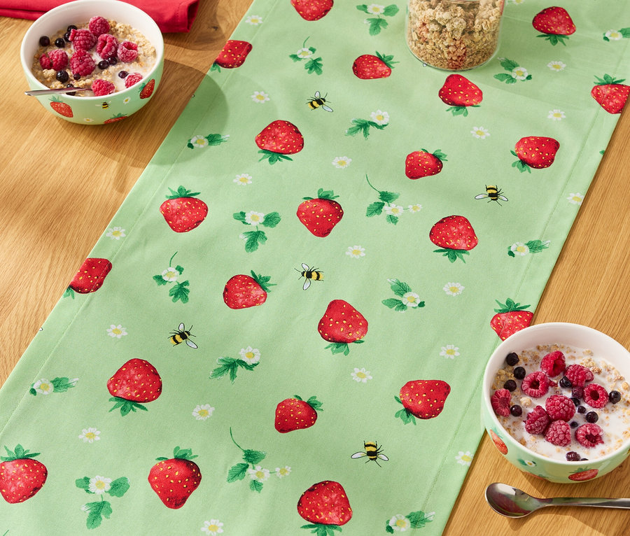 Auf einem Holztisch liegt ein Tischläufer mit Erdbeermuster, darauf Müslischalen, ein Glas Müsli und eine Milchflasche.