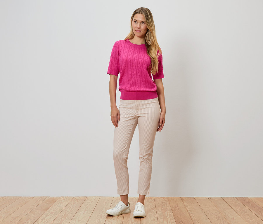 Frau steht vor einer Wand, trägt einen pinken Pullover, eine gestreifte Hose und weiße Schuhe.