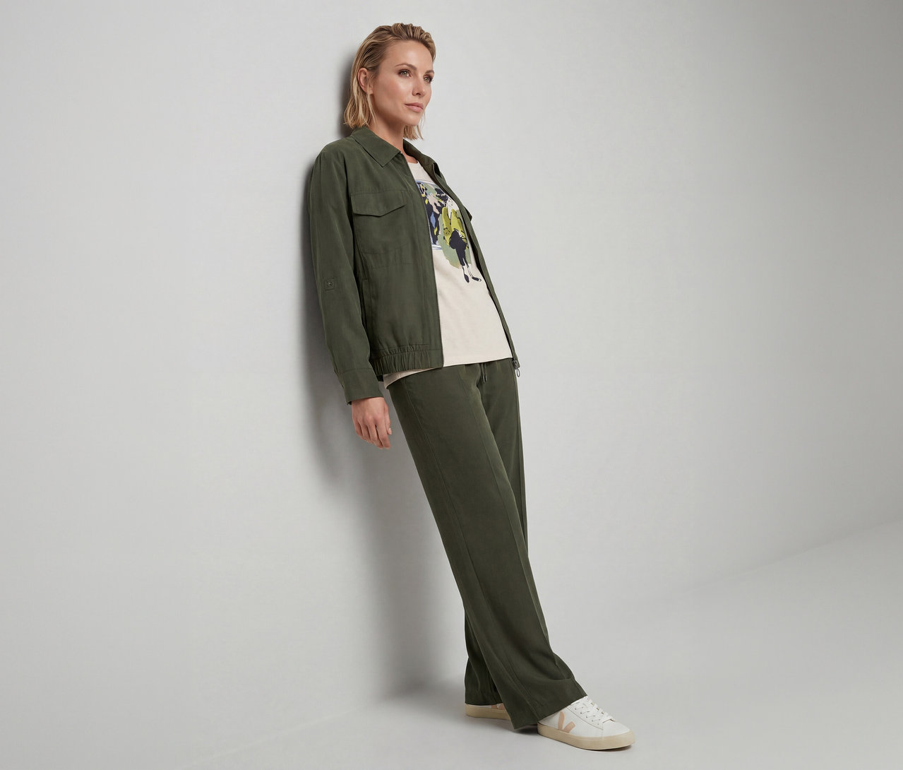 Frau mit kurzen blonden Haaren lehnt an einer Wand, trägt eine dunkelgrüne Jacke, ein helles T-Shirt mit Grafikprint, dunkelgrüne weite Hosen und weiße Sneaker.