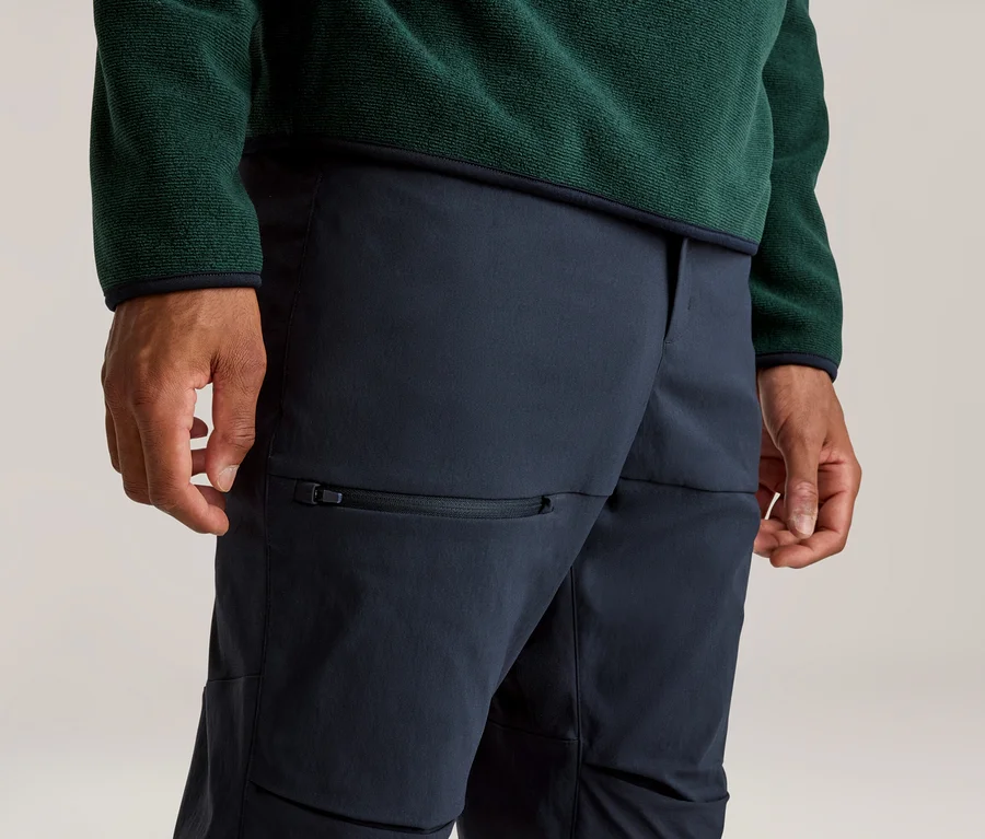 Nahaufnahme einer Person, die ein dunkelgrünes Fleece-Oberteil und eine dunkelblaue Hose mit Reißverschlusstasche am Oberschenkel trägt. Die Hände sind an den Seiten sichtbar.