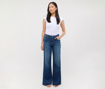 Eine Frau steht in einem weißen T-Shirt und einer blauen Jeans mit weitem Bein, die Hände in den Taschen.