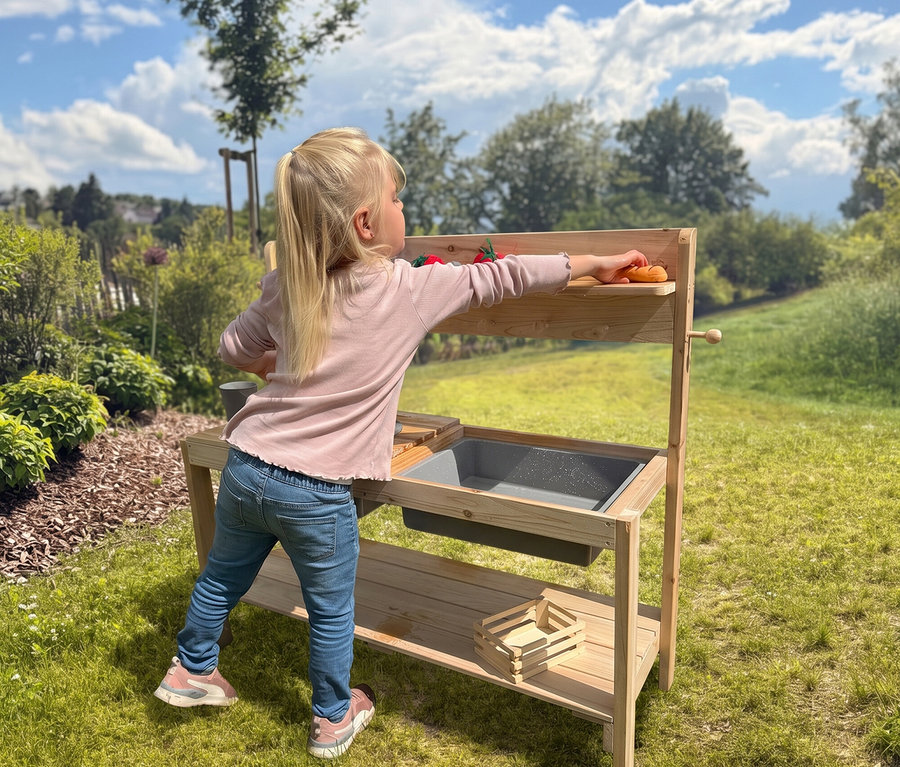 Blondes Mädchen spielt draußen mit einer Matschküche aus Holz und greift nach Spielzeuggemüse auf dem oberen Regal. Die Matschküche hat ein graues Waschbecken und eine Holzkiste auf dem unteren Regal, vor einem sonnigen, grasbewachsenen Hintergrund.