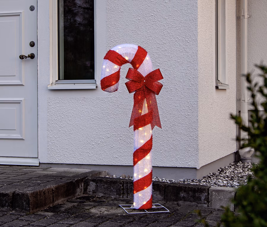 Große, beleuchtete Zuckerstangen-Dekoration mit rot-weißen Streifen und roter Schleife vor einem weißen Haus auf einem gepflasterten Weg.