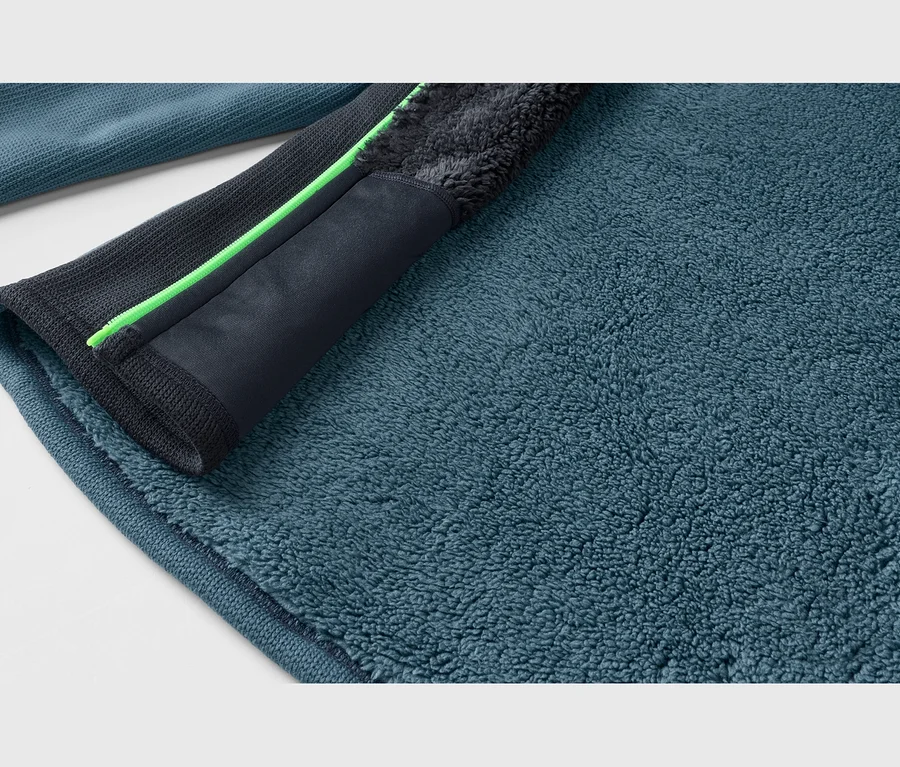 Nahaufnahme eines petrolfarbenen Fleece-Stoffs, der seine flauschige Textur zeigt. Ein dunkelgrauer Besatz mit einem neongrünen Reißverschlussdetail verläuft diagonal über den Stoff.