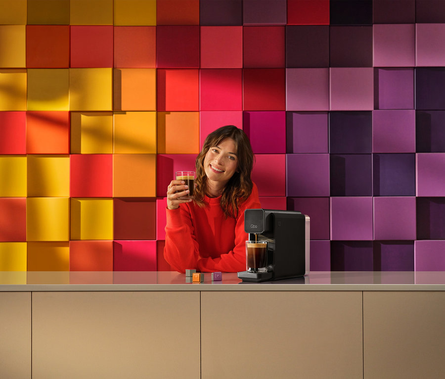 Lächelnde Frau in rotem Pullover hält ein Glas Kaffee, während eine schwarze »Qbo ONE« Kapselmaschine Kaffee in ein Glas füllt. Bunte Kapseln liegen auf der Theke.