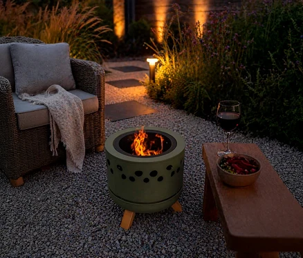 Abendliche Gartenszene mit einem brennenden Feuerkorb in Olivgrün, einem Korbsessel mit Decke und einer Holzbank mit Rotwein und Snacks auf Kiesboden.