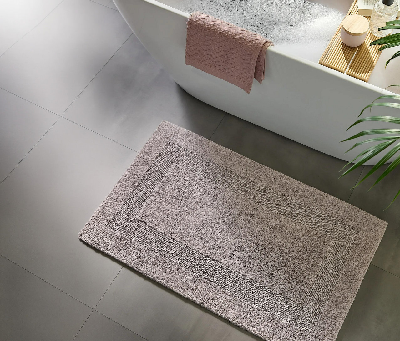 Heller Badteppich mit Randmuster auf grauem Fliesenboden neben einer weißen Badewanne mit Schaum, einem rosa Handtuch und einem Holztablett mit Kerze und Flasche.