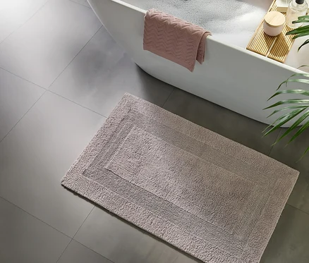 Heller Badteppich mit Randmuster auf grauem Fliesenboden neben einer weißen Badewanne mit Schaum, einem rosa Handtuch und einem Holztablett mit Kerze und Flasche.