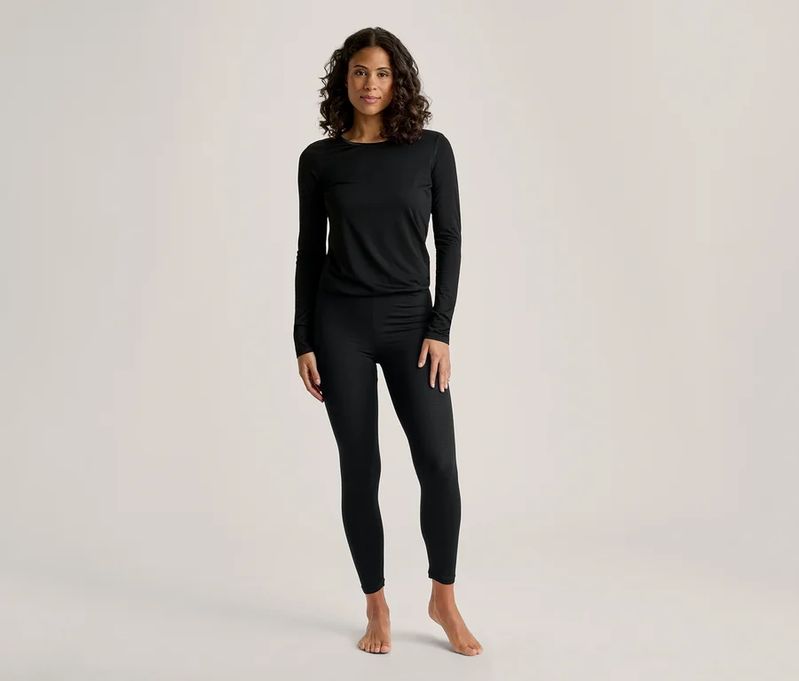 Eine Frau mit dunklem, lockigem Haar steht frontal und trägt ein schwarzes Langarmshirt und schwarze Leggings. Sie ist barfuß.